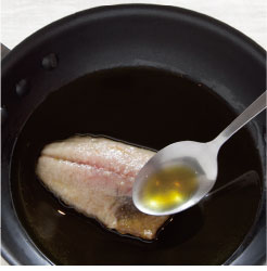 フライパンの魚にオリーブオイルをかけ皮目を焼き上げている調理写真